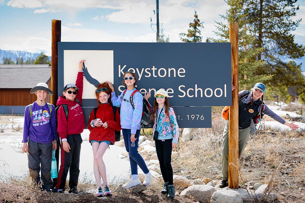 Field trip students and a KSS instructor smile in front of the entrance sign to campus.