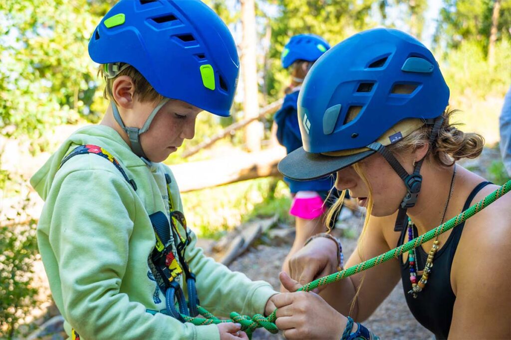 A camp counselor shows a camper how to tie their harness in for rock climbing.